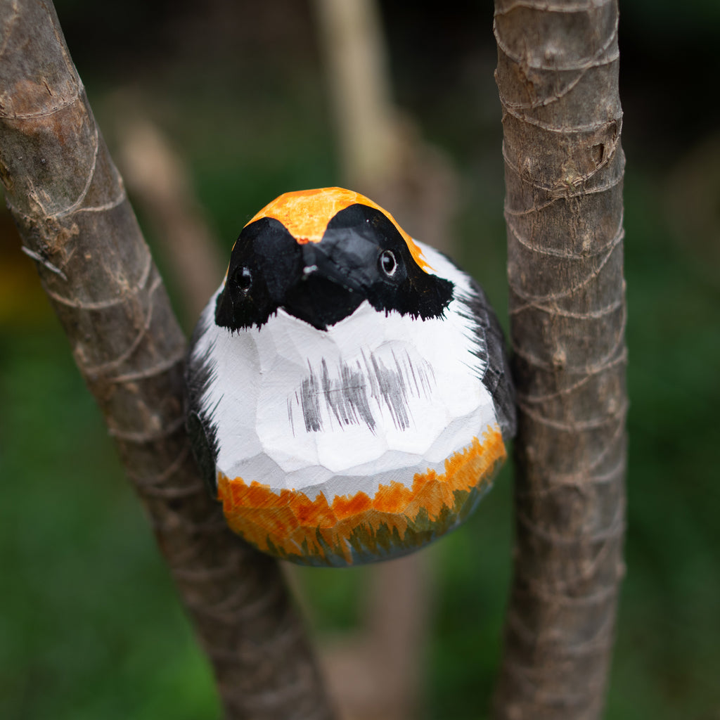 A hand-carved wooden figurine of a Black-throated Bushtit perched on a branch, with detailed black, white, and orange markings.