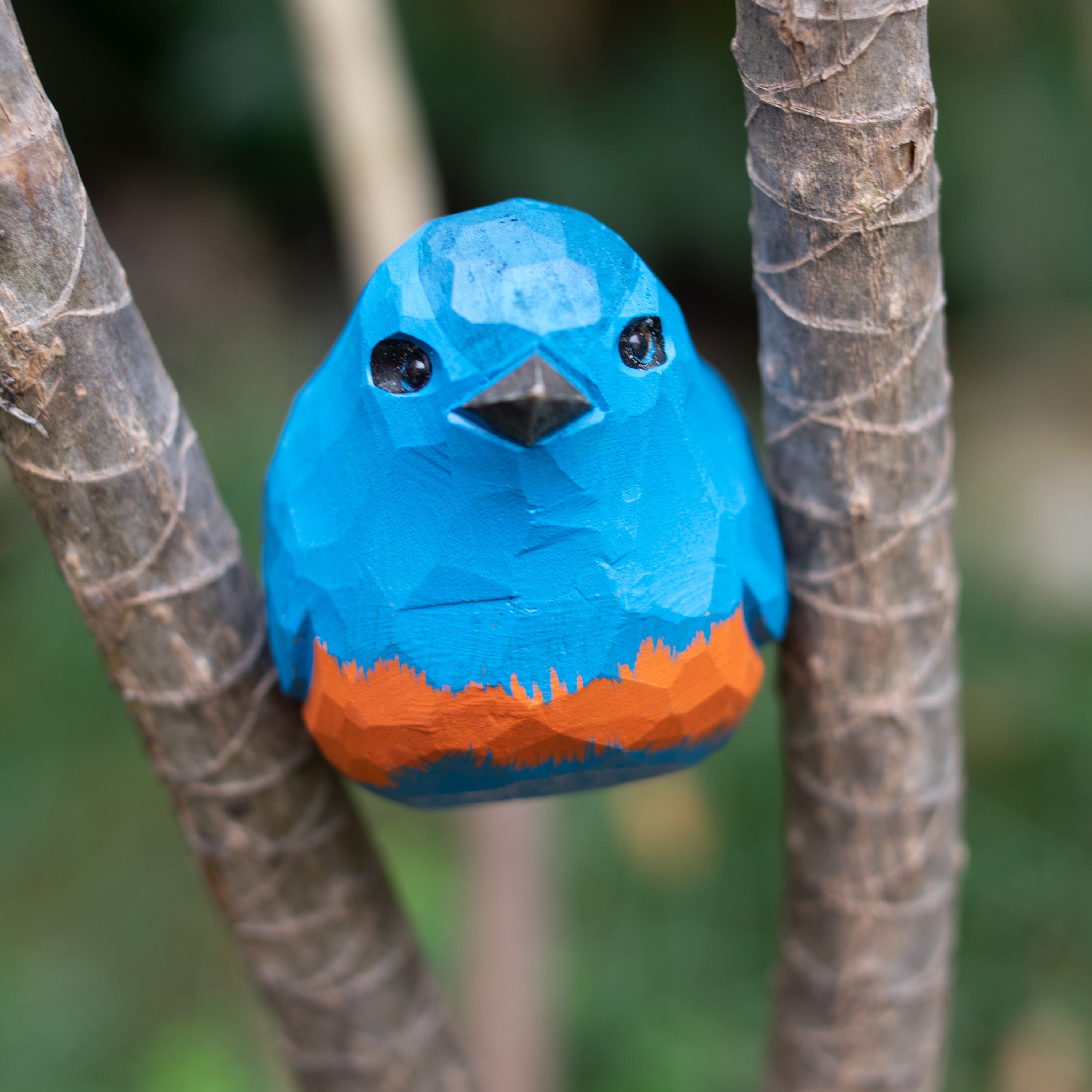 A hand-carved wooden figurine of a Blue-throated Flycatcher perched on a branch, with vibrant blue and orange colors.