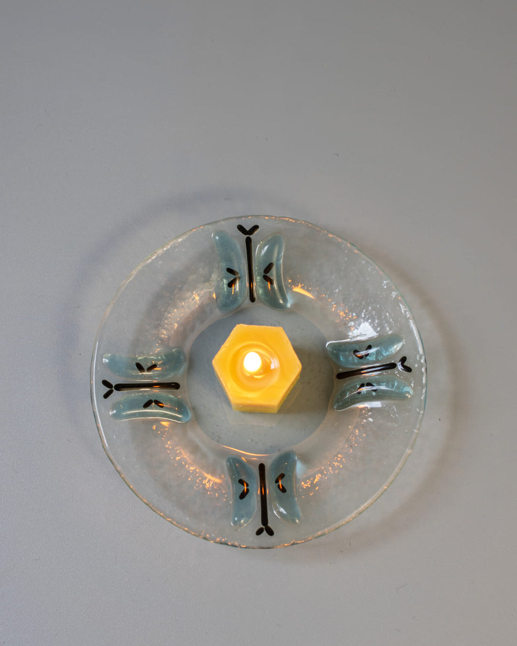 A 90s-style square fused glass taper candle holder with a blue butterfly motif, holding a lit yellow beeswax candle against a gray background.