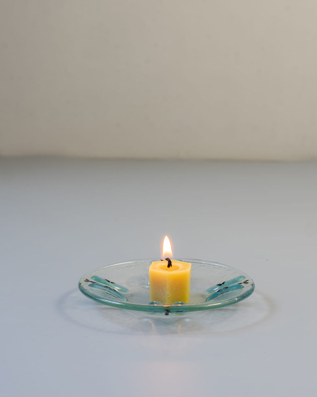 A 90s-style square fused glass taper candle holder with a blue butterfly motif, holding a lit yellow beeswax candle against a gray background.