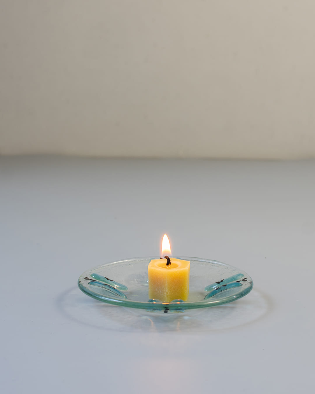 A 90s-style square fused glass taper candle holder with a blue butterfly motif, holding a lit yellow beeswax candle against a gray background.