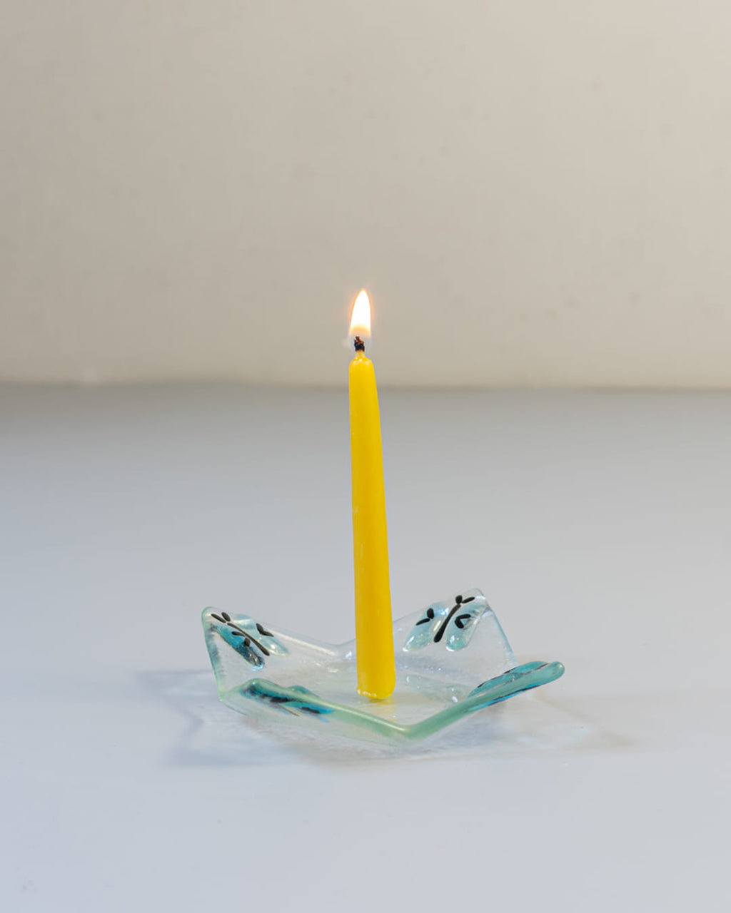 A 90s-style square fused glass taper candle holder with a blue butterfly motif, holding a lit yellow beeswax candle against a gray background.