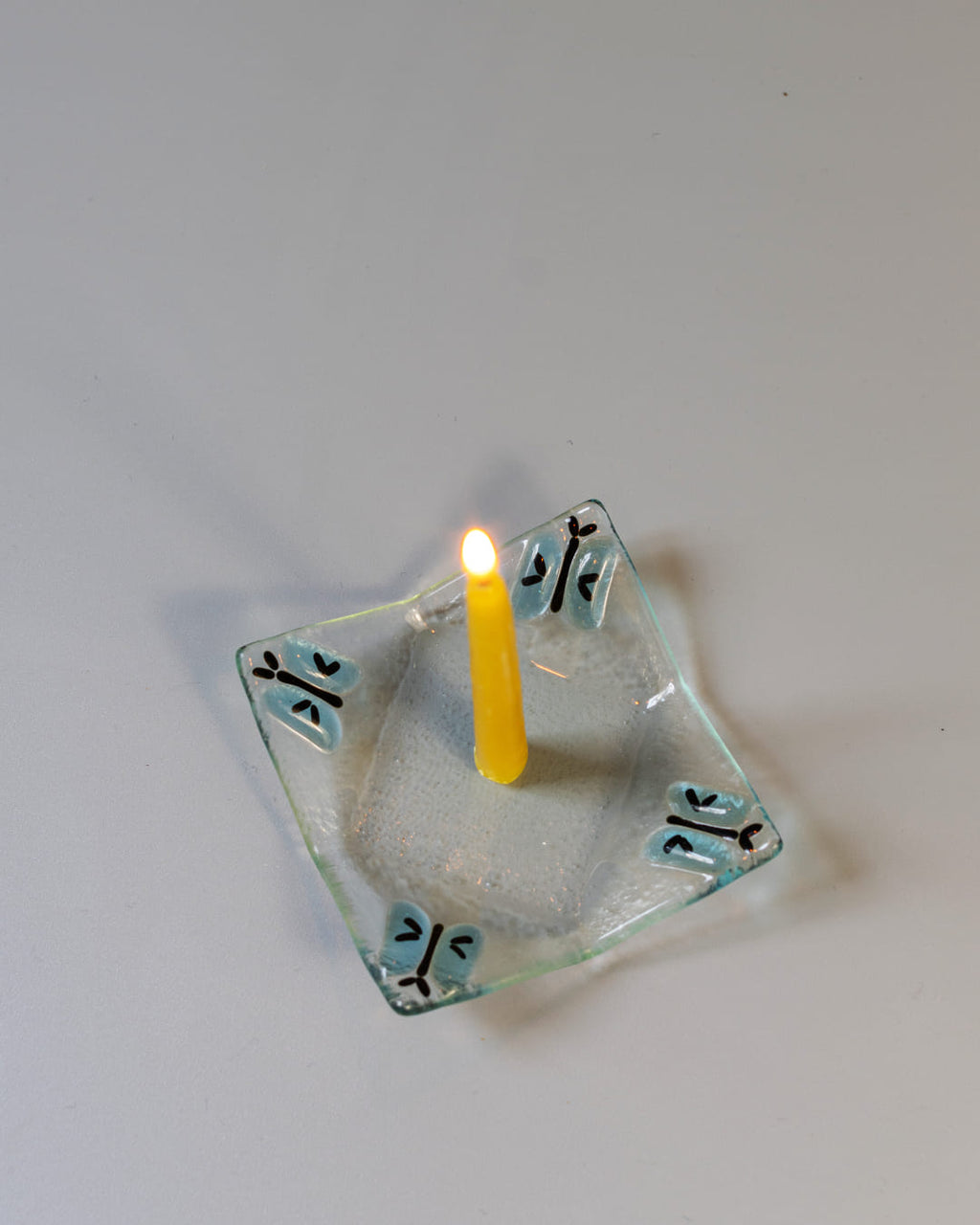 A 90s-style square fused glass taper candle holder with a blue butterfly motif, holding a lit yellow beeswax candle against a gray background.