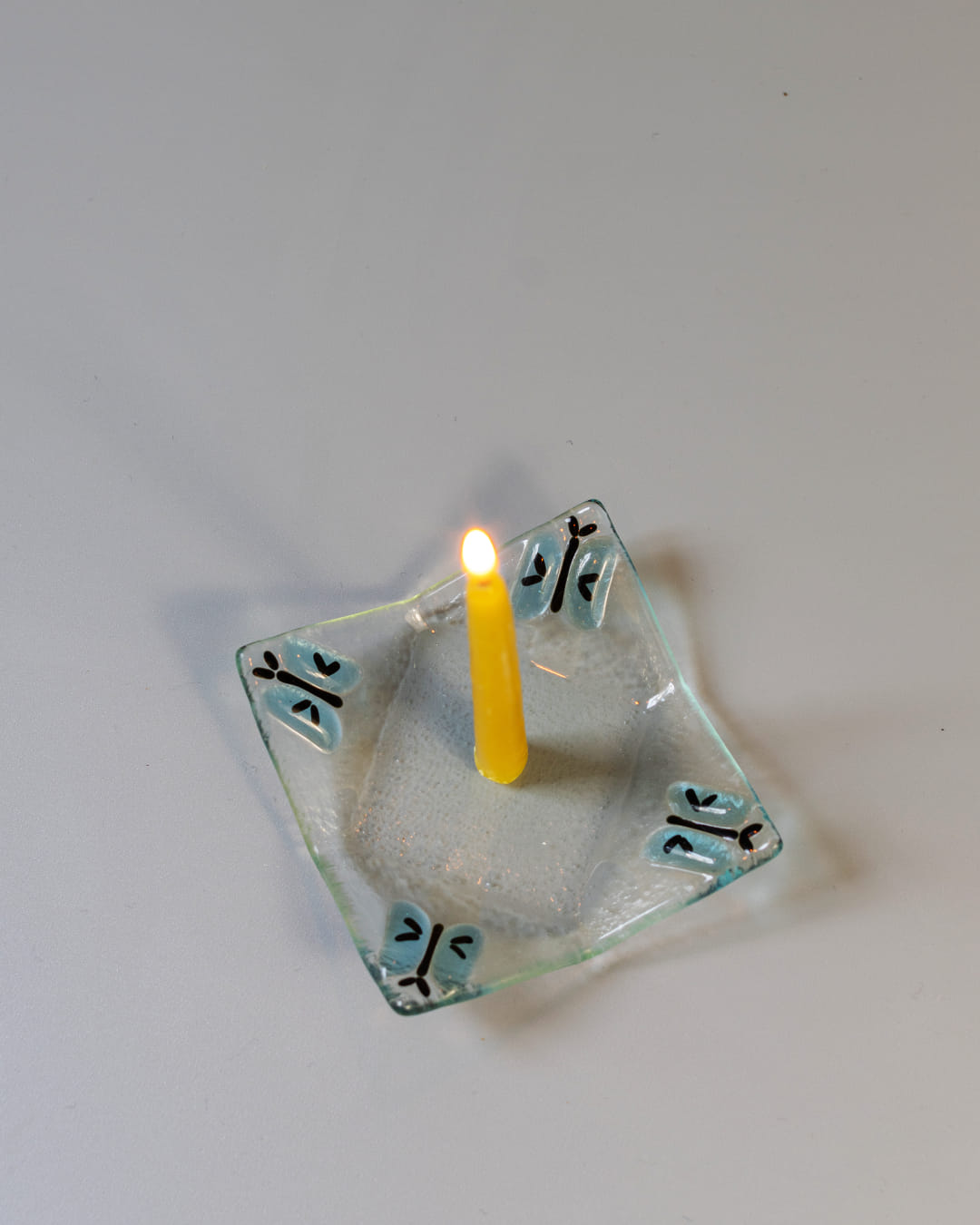 A 90s-style square fused glass taper candle holder with a blue butterfly motif, holding a lit yellow beeswax candle against a gray background.