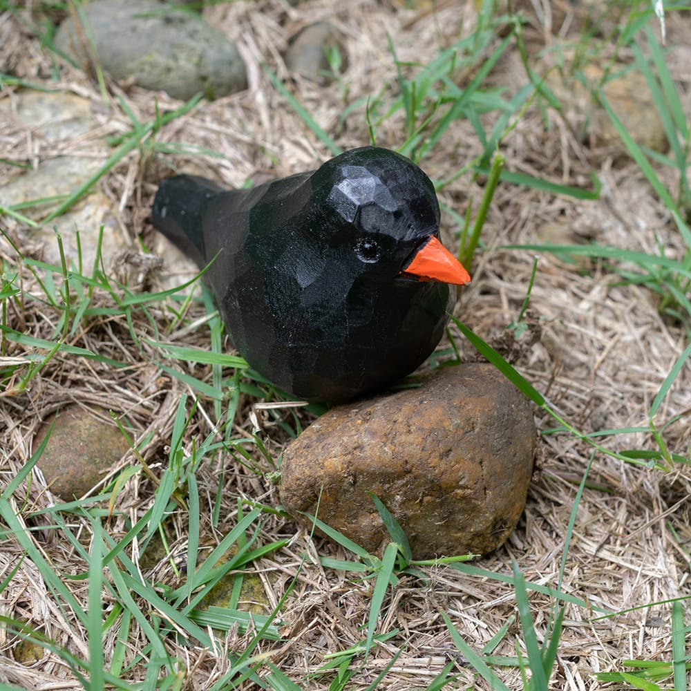 Wooden blackbird figurine with a red beak on grass and rocks