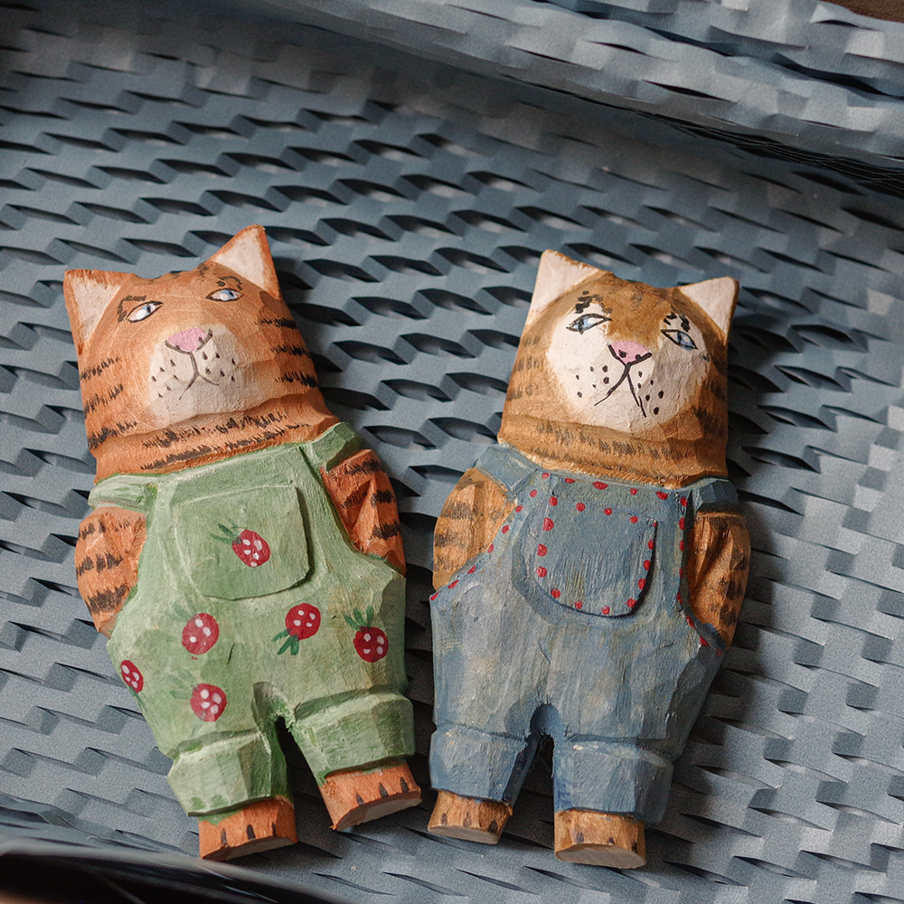 Two wooden cat magnets wearing overalls on a textured surface