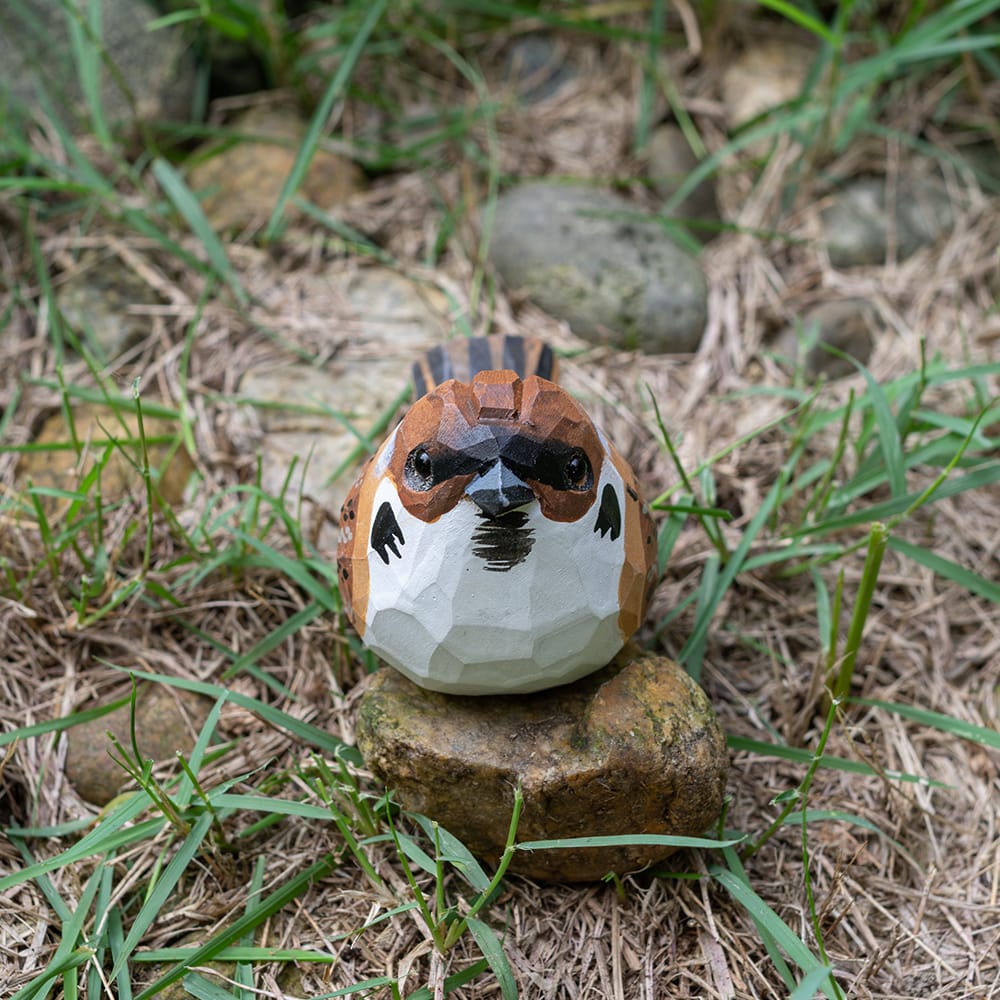 Eurasian Tree Sparrow sculpture on a stone in a natural setting with grass and rocks.