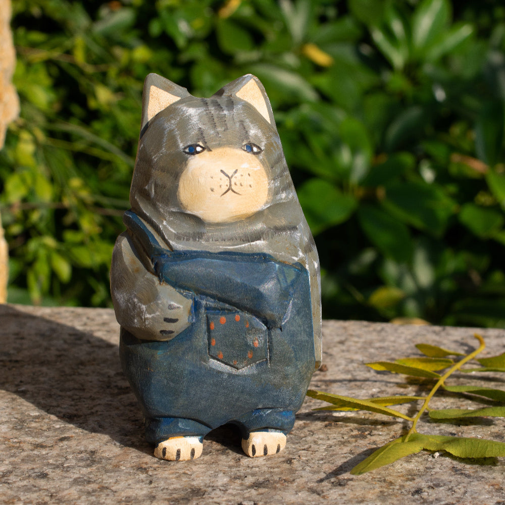 A wooden figurine of a gray tabby cat wearing overalls, positioned outdoors with foliage in the background.