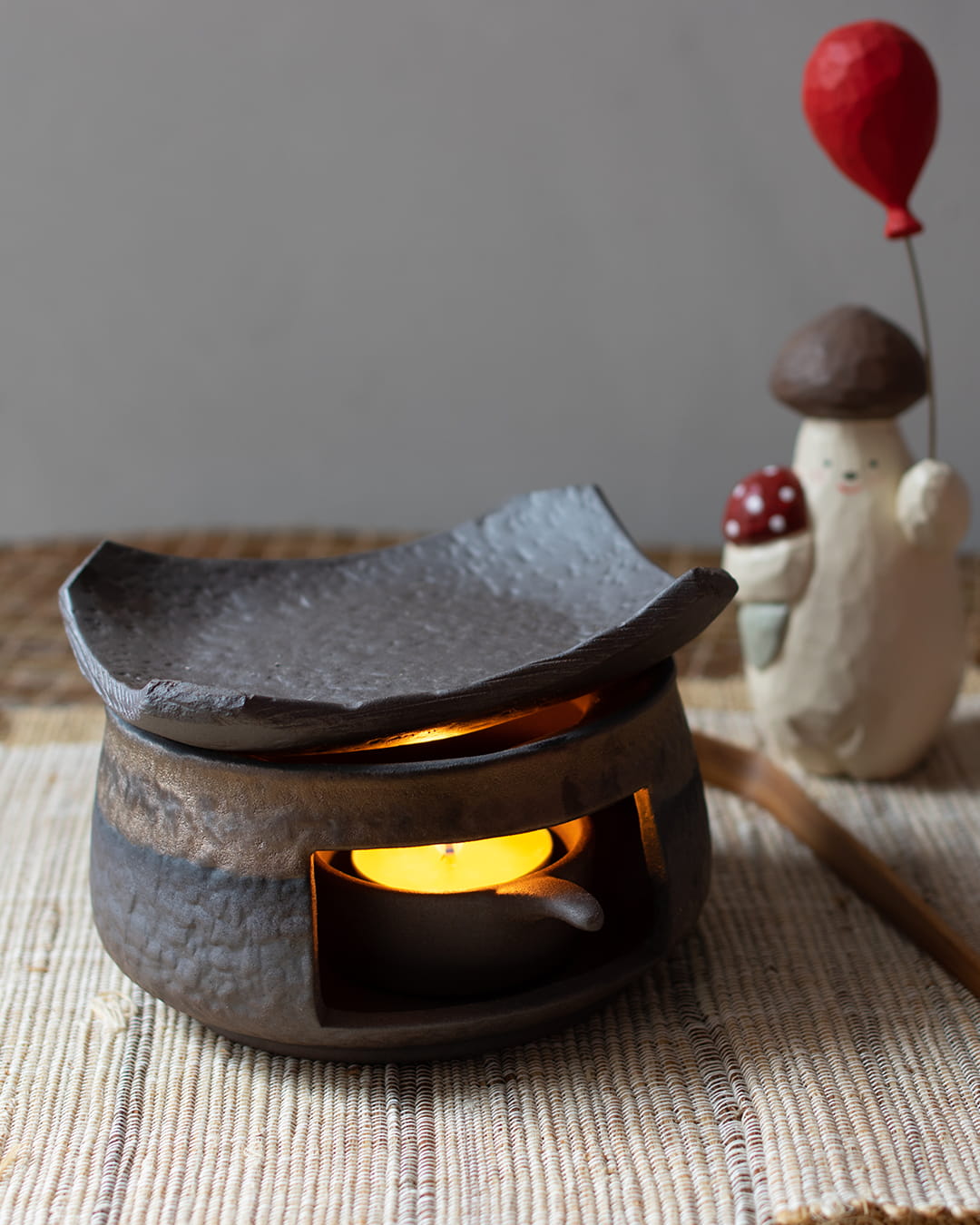 A dark, stone-textured ceramic fragrance warmer (Kawa) with a lit tealight inside, topped with decorative items in the background.