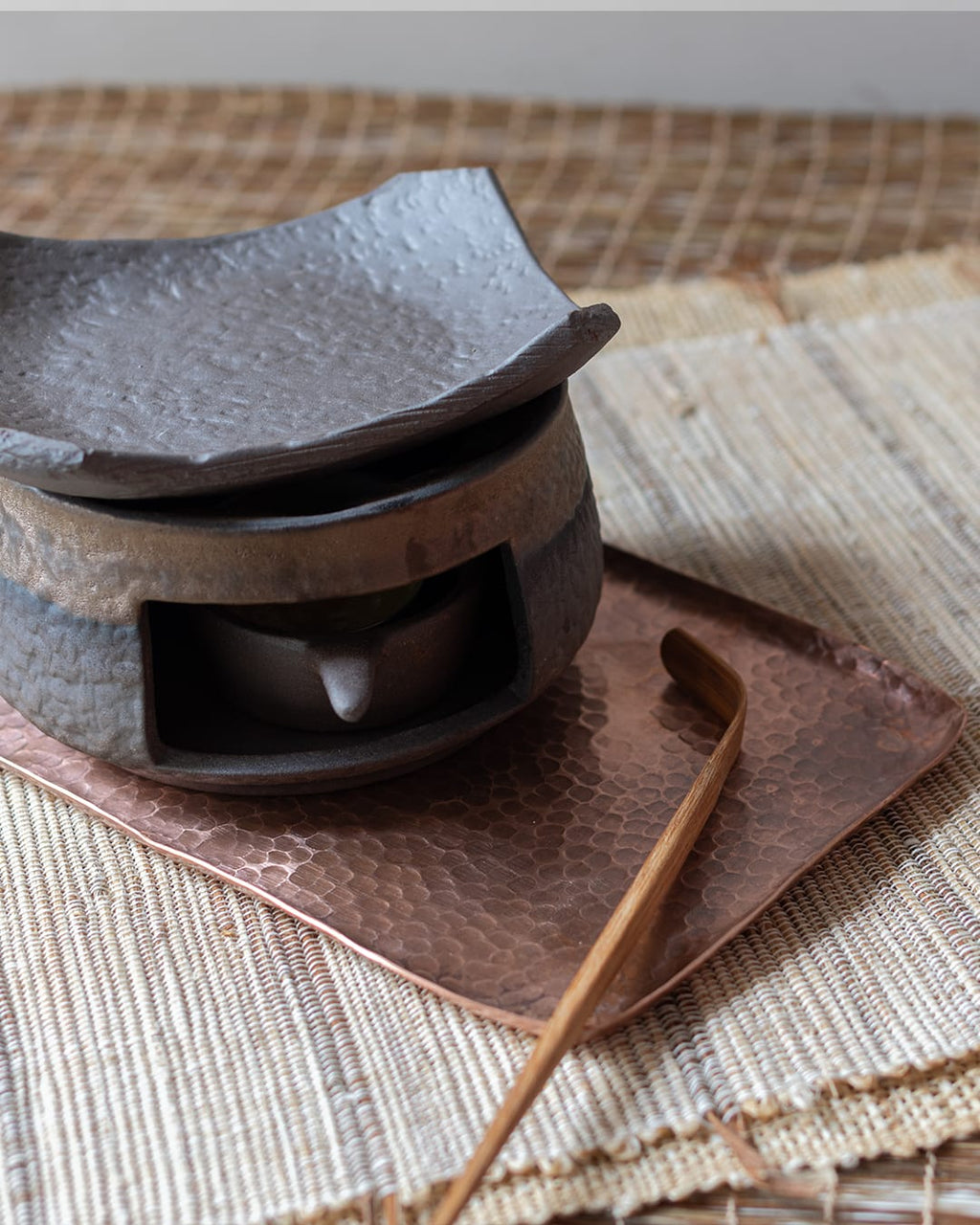 A dark, stone-textured ceramic fragrance warmer (Kawa) with a lit tealight inside, topped with a traditional Japanese-style tile, sitting on a copper plate.