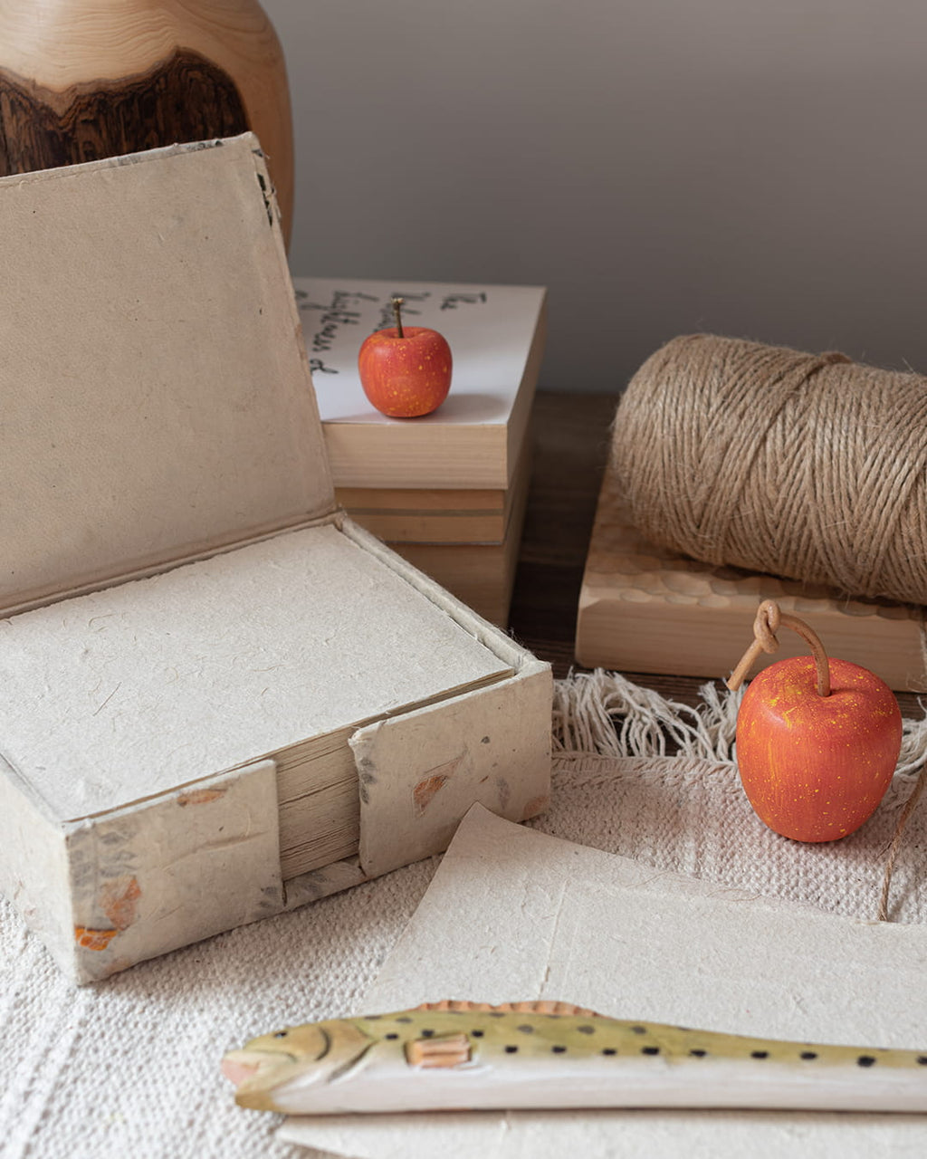 A handmade Lokta paper box and two note sheets, with a hand-carved wooden apple ornament resting on top of the paper, on a linen table sheet.