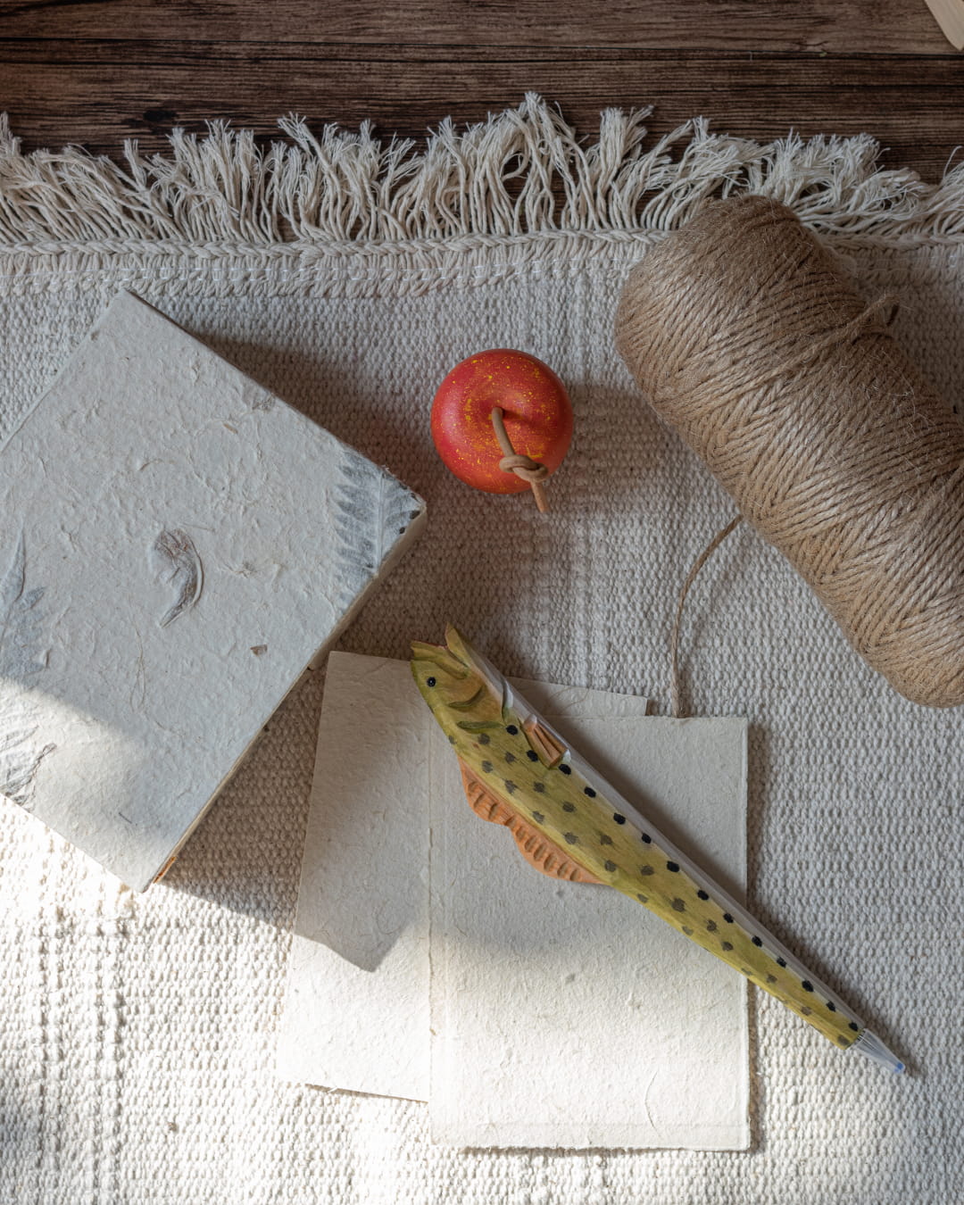 A handmade Lokta paper box and two note sheets, with a hand-carved wooden apple ornament and handcrafted fish pen resting on top of the paper, on a linen table sheet.