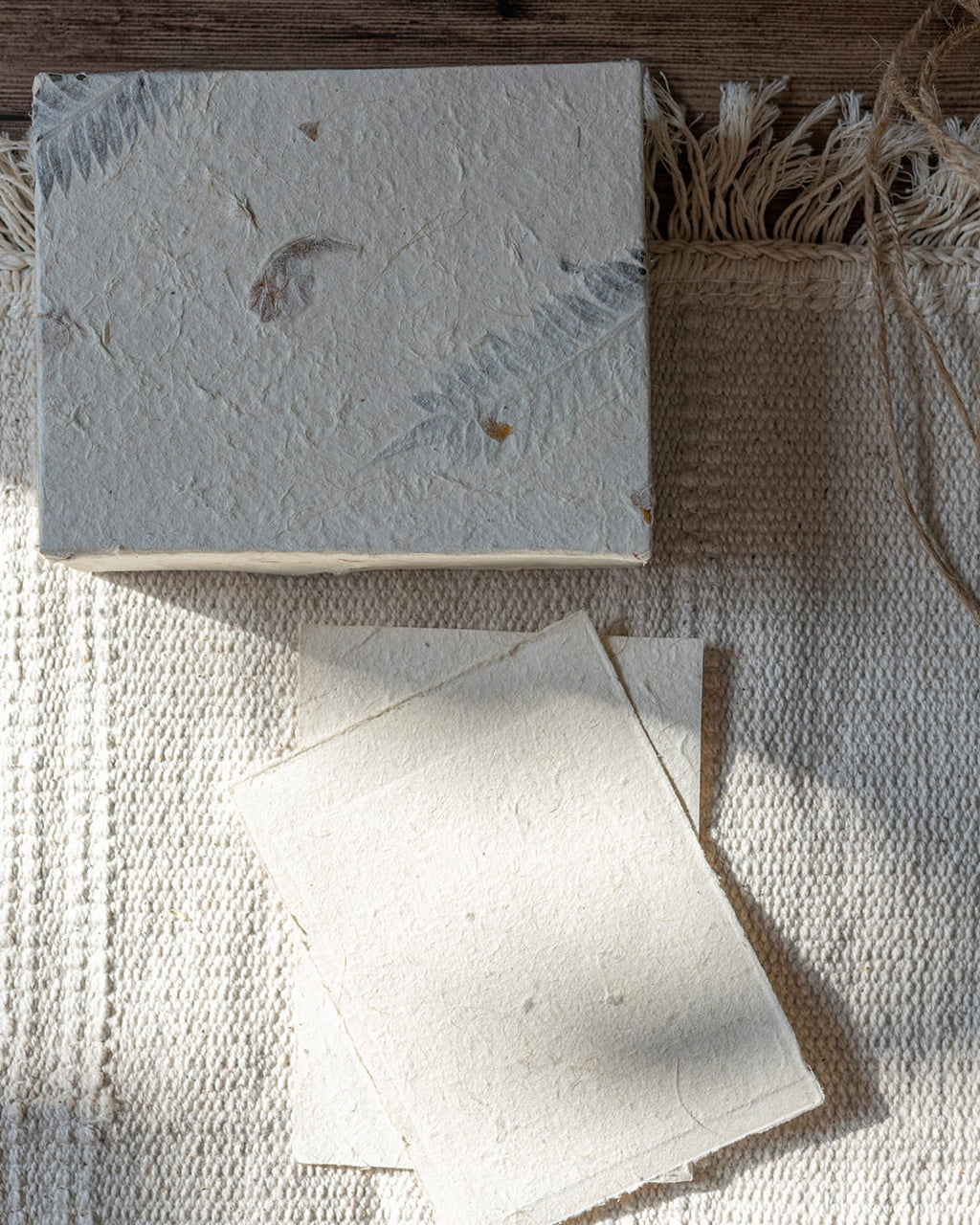 A handmade Lokta paper box and two note sheets, with a hand-carved mackerel fish pen resting on top of the paper, on a dark wood table.