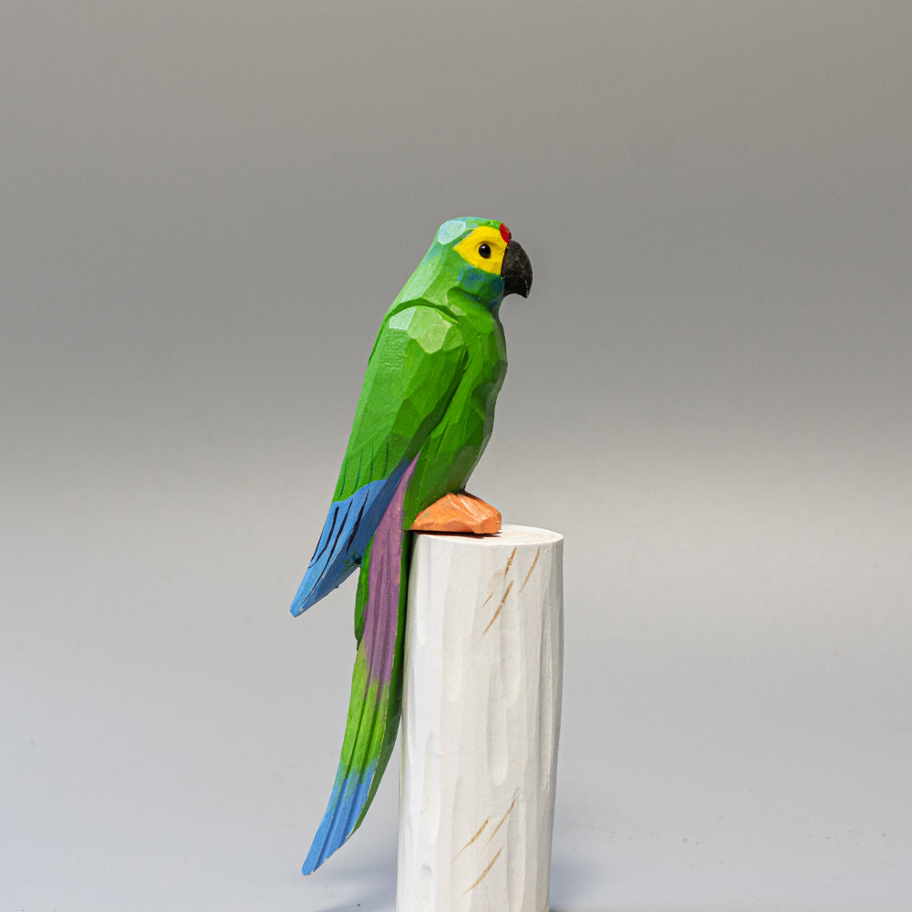 Hand-carved wooden Military Macaw figurine, green with blue wing accents and long tail, on a white cylindrical stand. 