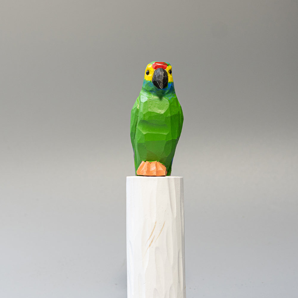 Hand-carved wooden Military Macaw figurine, green with blue wing accents and long tail, on a white cylindrical stand. 