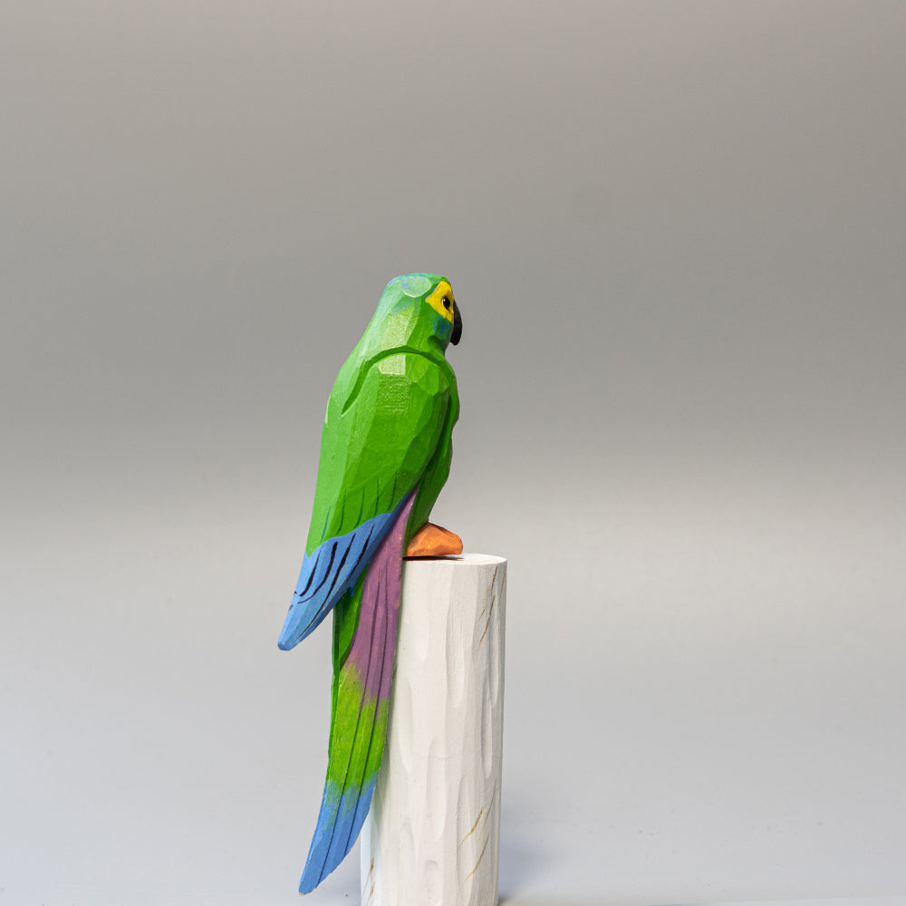 Hand-carved wooden Military Macaw figurine, green with blue wing accents and long tail, on a white cylindrical stand. 