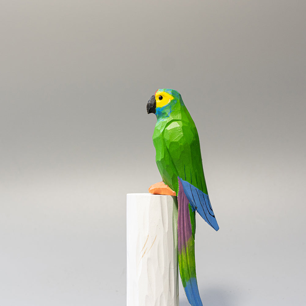 Hand-carved wooden Military Macaw figurine, green with blue wing accents and long tail, on a white cylindrical stand. 