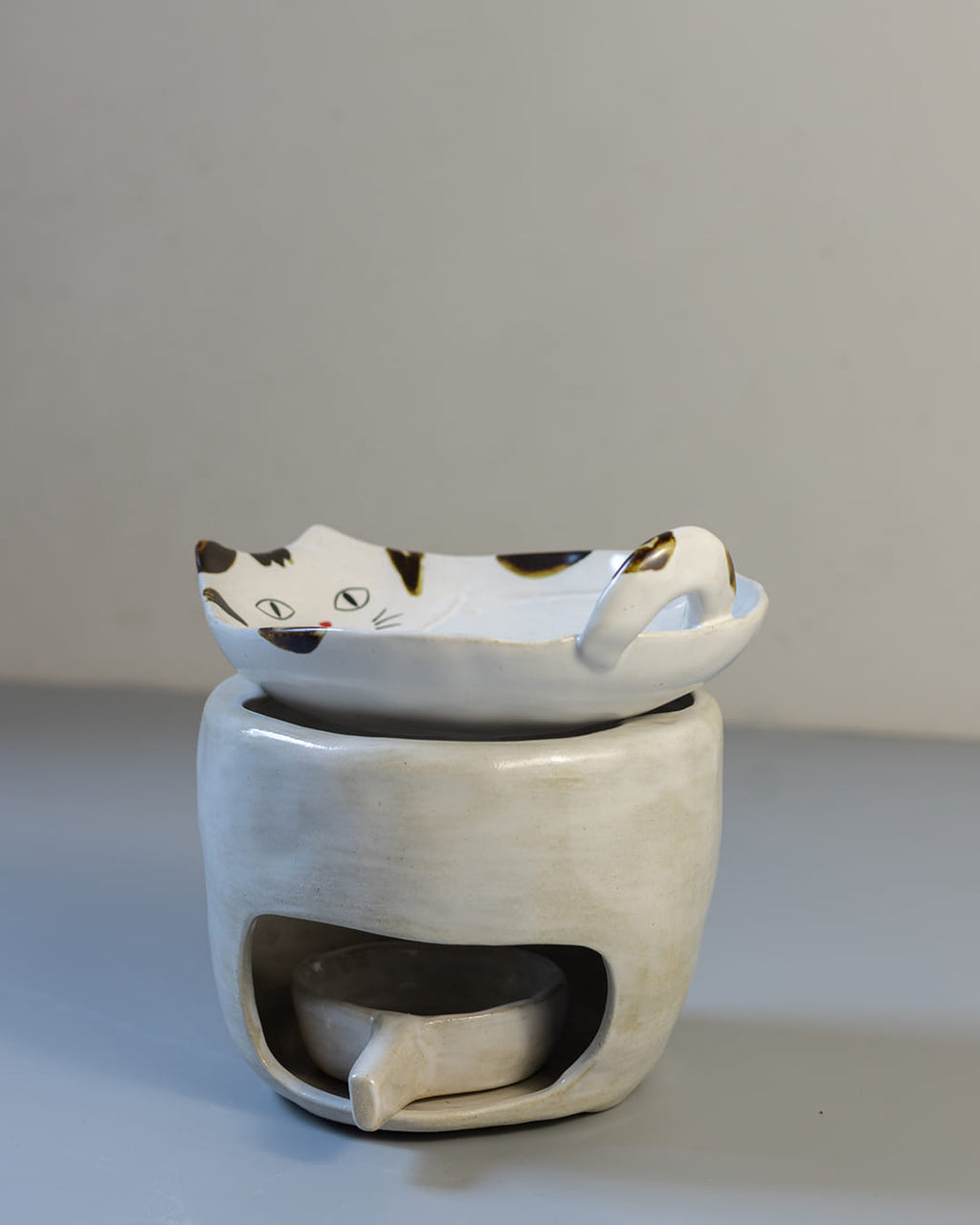 A cute, cat-shaped ceramic fragrance warmer with a white Kohiki finish, resting on its stove base with a lit tealight visible inside.