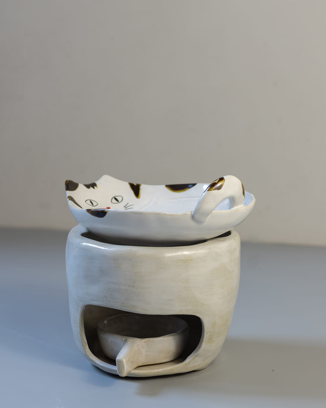 A cute, cat-shaped ceramic fragrance warmer with a white Kohiki finish, resting on its stove base with a lit tealight visible inside.