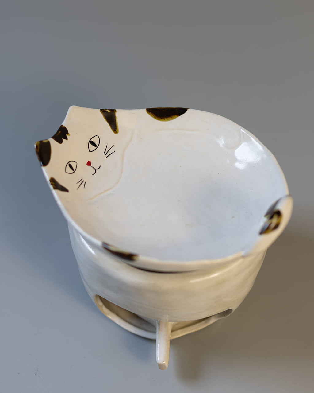 A cute, cat-shaped ceramic fragrance warmer with a white Kohiki finish, resting on its stove base with a lit tealight visible inside.