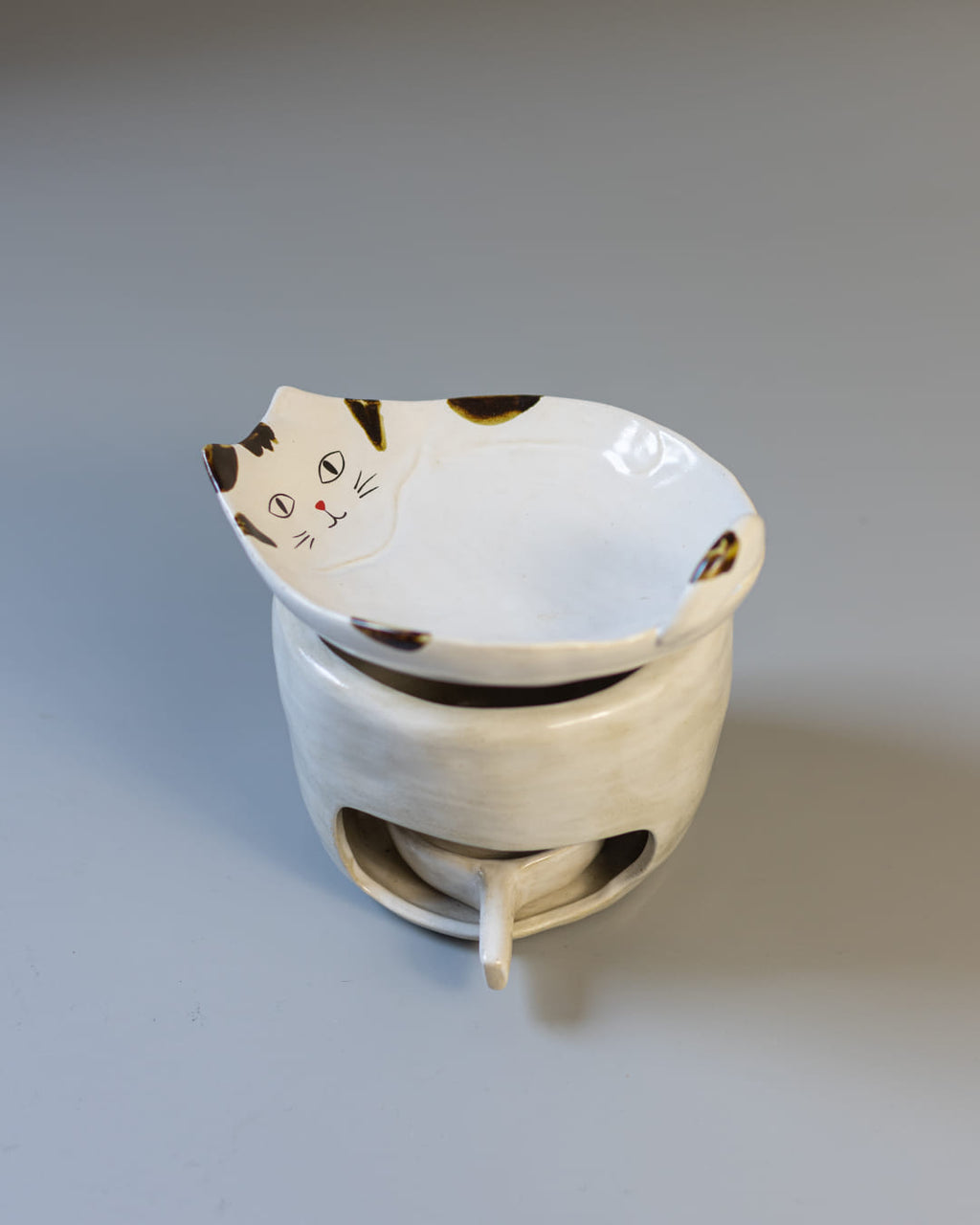 A cute, cat-shaped ceramic fragrance warmer with a white Kohiki finish, resting on its stove base with a lit tealight visible inside.