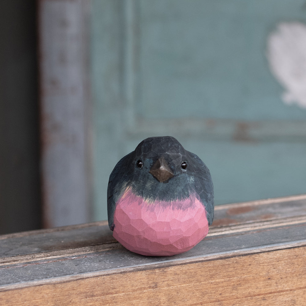 A hand-carved wooden figurine of a Pink Robin with a soft pink chest and black feathers, perched on a wooden surface.