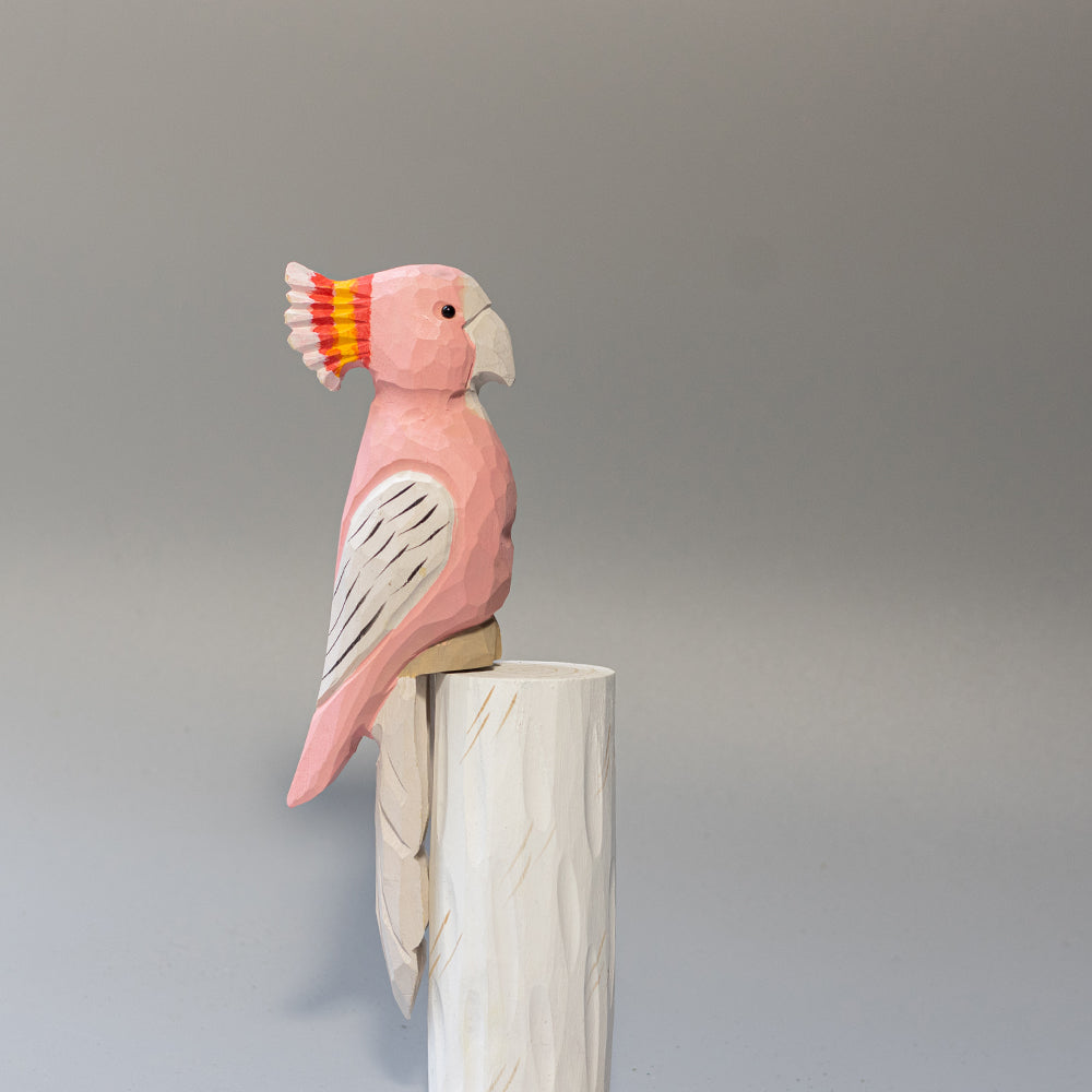 Hand-carved wooden Major Mitchell's Cockatoo figurine with pink body and vibrant red-yellow crest, on a white cylindrical stand. 