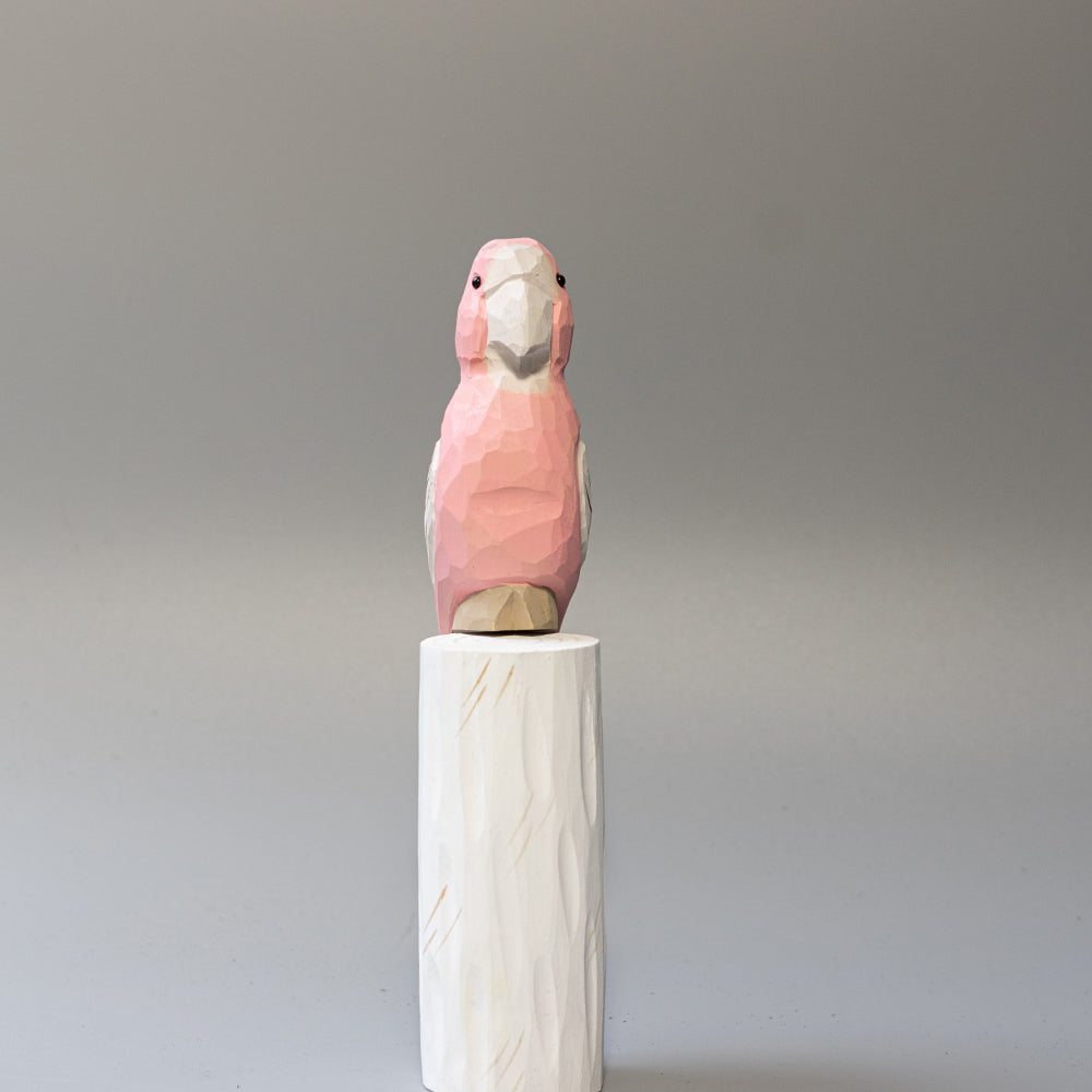 Hand-carved wooden Major Mitchell's Cockatoo figurine with pink body and vibrant red-yellow crest, on a white cylindrical stand. 