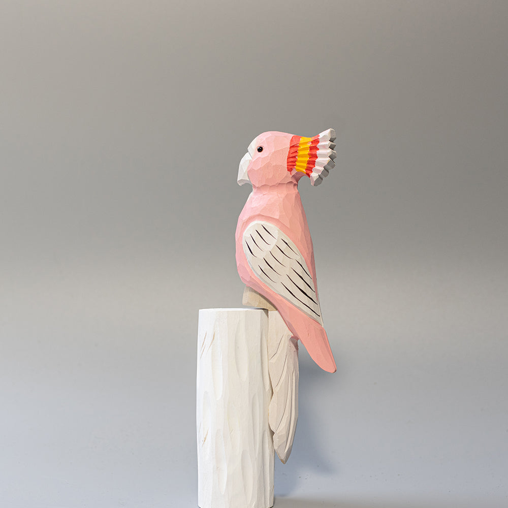 Hand-carved wooden Major Mitchell's Cockatoo figurine with pink body and vibrant red-yellow crest, on a white cylindrical stand. 