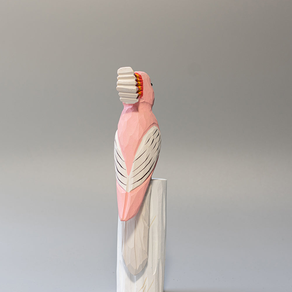 Hand-carved wooden Major Mitchell's Cockatoo figurine with pink body and vibrant red-yellow crest, on a white cylindrical stand. 