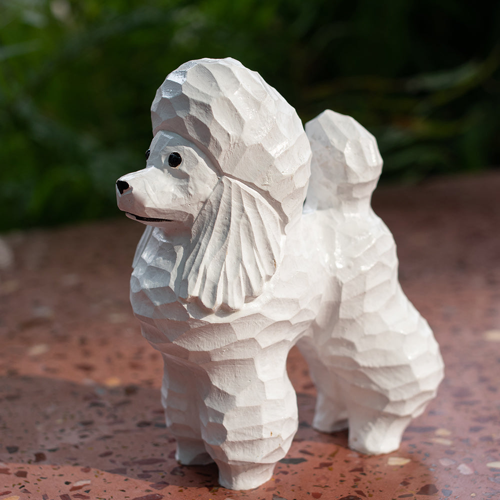 A hand-carved basswood figurine of a white poodle with detailed carving and polished wood grain, standing on a flat surface.