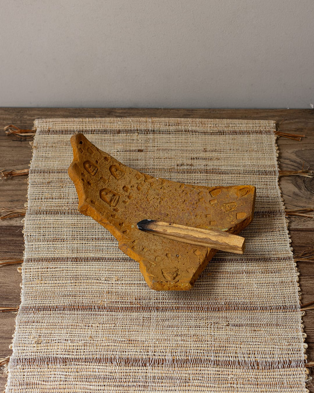 An earthy yellow cement incense plate, shaped like an ancient fragment, holding both a smoldering palo santo stick and a bundle of white sage.