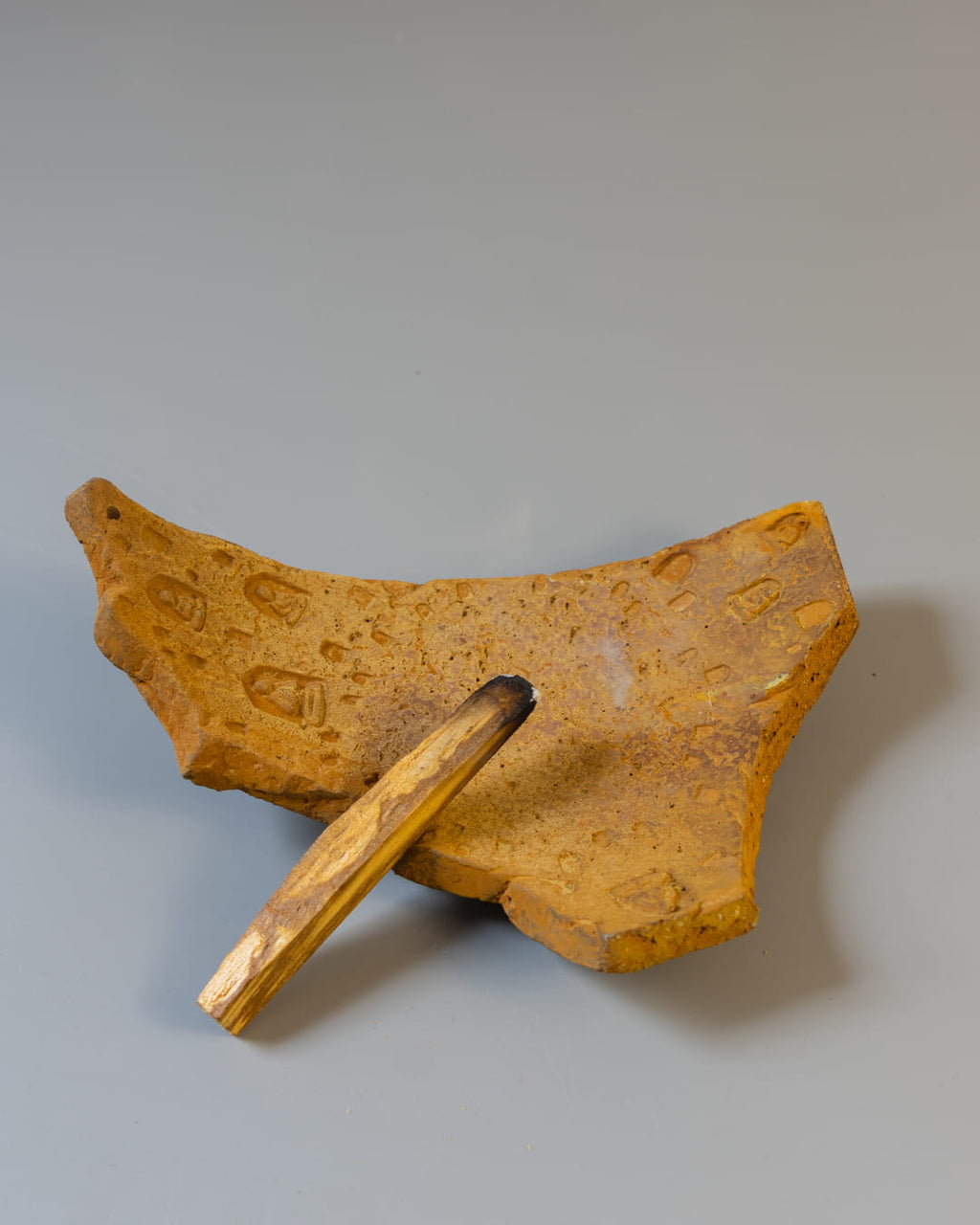 An earthy yellow cement incense plate, shaped like an ancient fragment, holding both a smoldering palo santo stick and a bundle of white sage.