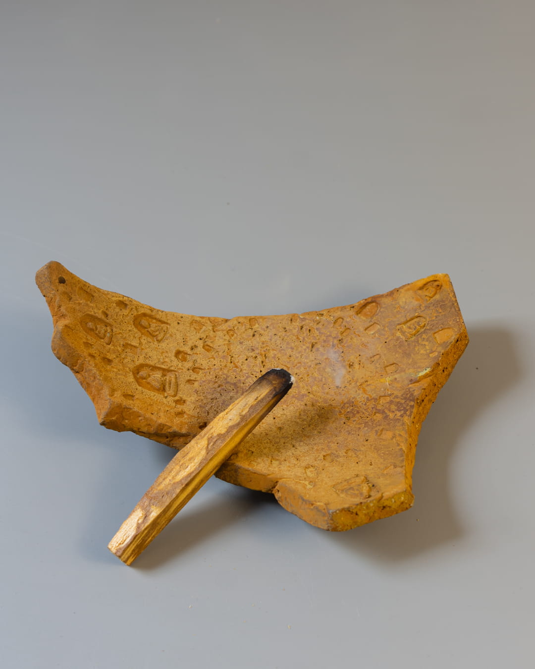 An earthy yellow cement incense plate, shaped like an ancient fragment, holding both a smoldering palo santo stick and a bundle of white sage.