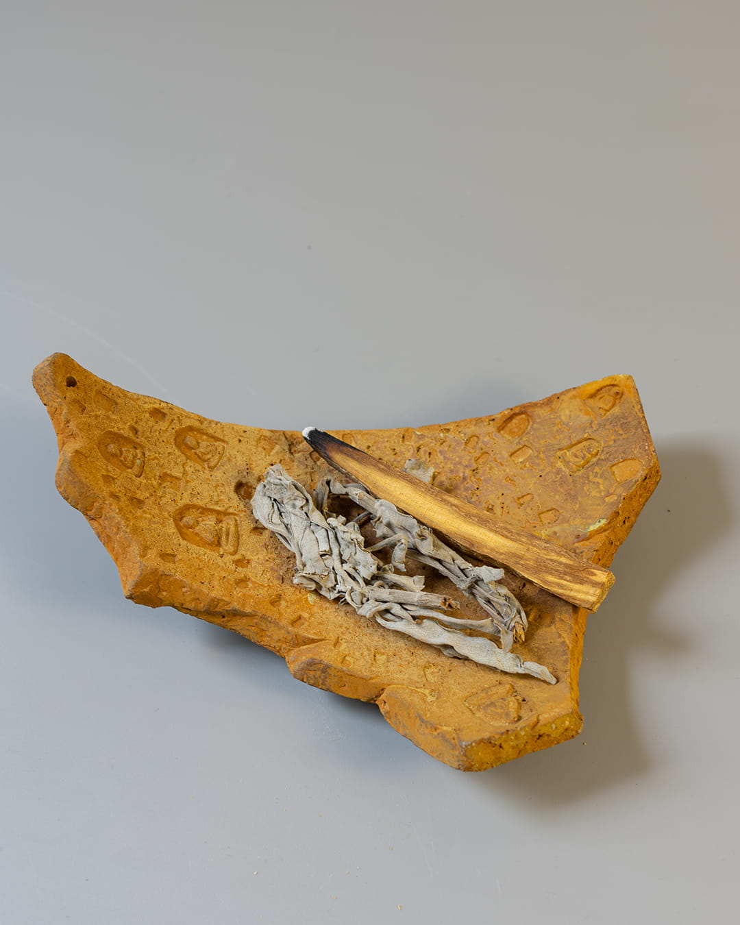 An earthy yellow cement incense plate, shaped like an ancient fragment, holding both a smoldering palo santo stick and a bundle of white sage.