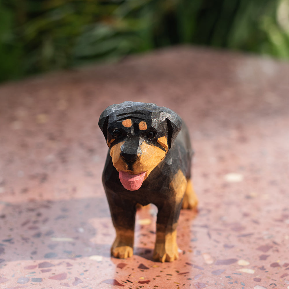 A hand-carved basswood figurine of a Rottweiler dog, with a black and brown color scheme, displayed on a flat surface with a blurred green background.