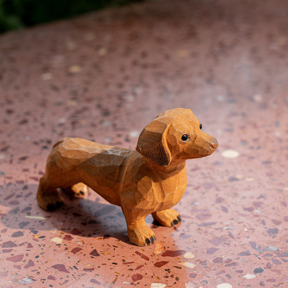 A wooden figurine of a tan dachshund dog, placed on a paved surface with a blurred background.