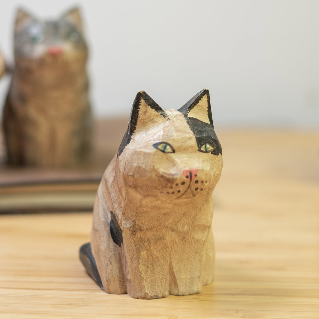 A hand-carved wooden figurine of a calico cat with a vintage finish, sitting  on a wooden surface.