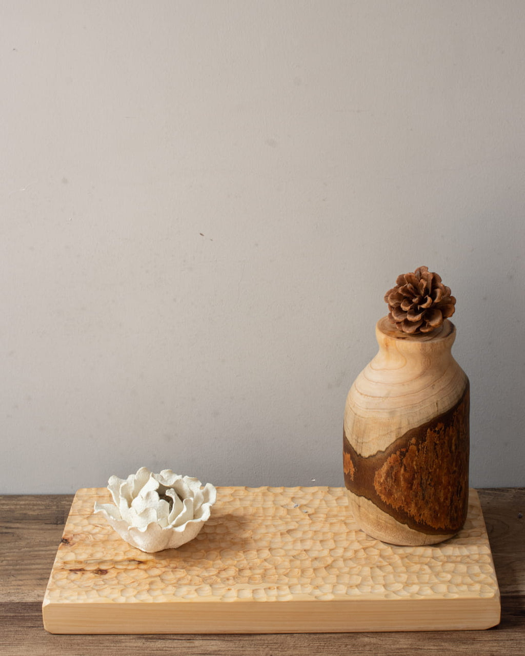 Wooden vase with a pine cone on top, white ceramic dish, and wooden tray against a gray background