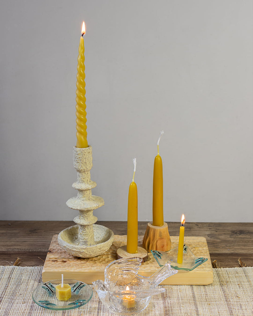 Beeswax candlestick with a lit candle on a wooden surface, surrounded by additional candles and a small dish.