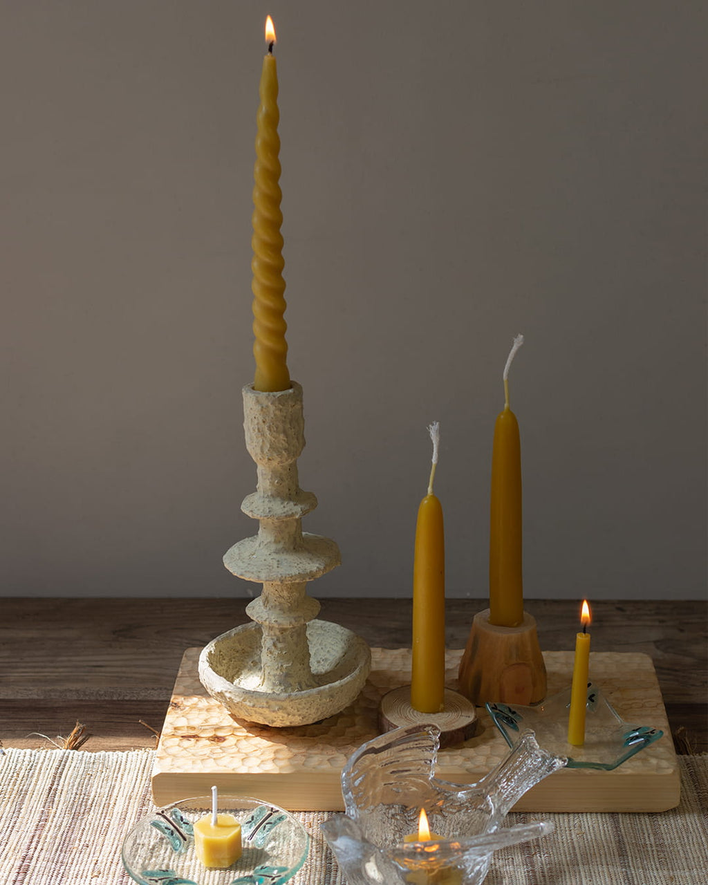 Beeswax candlestick with a lit candle on a wooden surface, surrounded by additional candles and a small dish.
