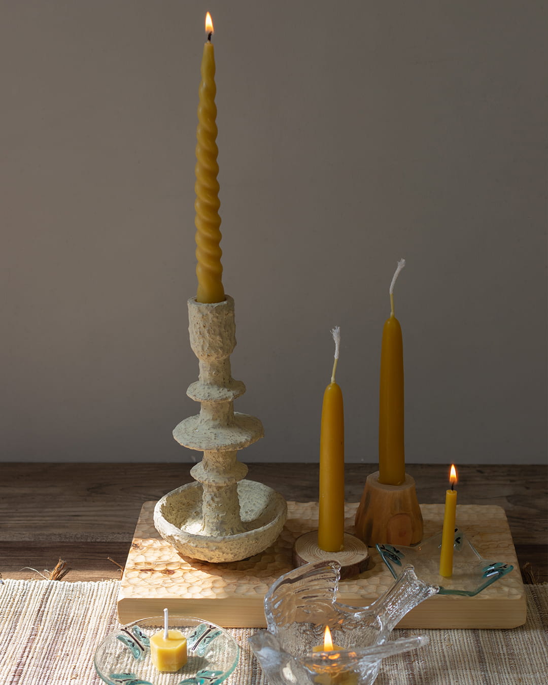 Beeswax candlestick with a lit candle on a wooden surface, surrounded by additional candles and a small dish.
