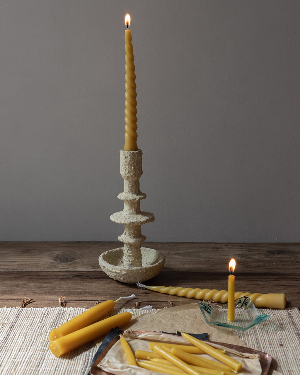 Beeswax candlestick with a lit candle on a wooden surface, surrounded by additional candles and a small dish.