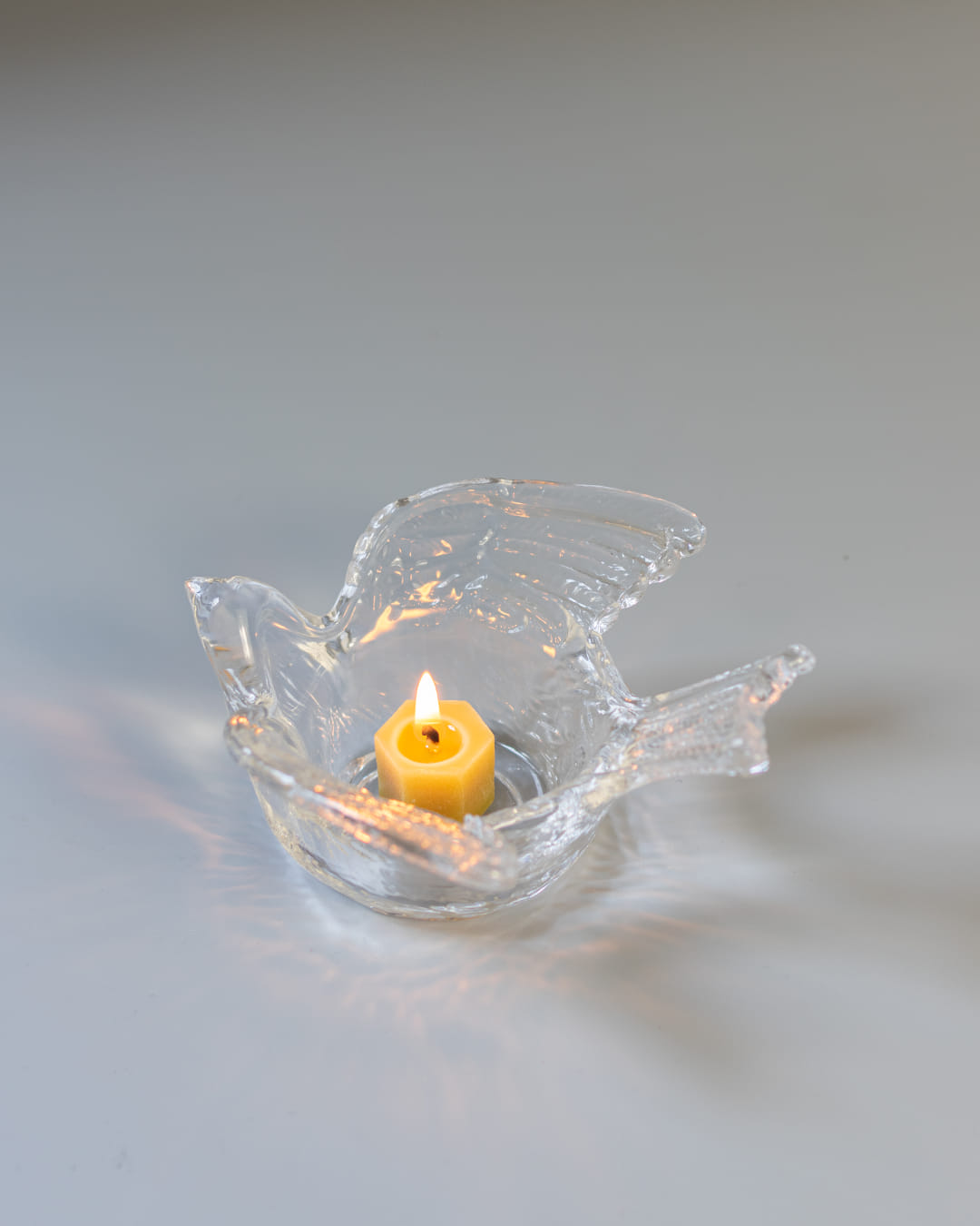 A clear, textured glass tealight holder shaped like a swallow, with a lit yellow tealight candle casting soft light patterns around it.