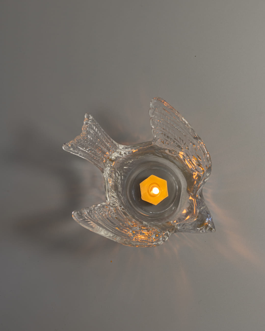 A clear, textured glass tealight holder shaped like a swallow, with a lit yellow tealight candle casting soft light patterns around it.