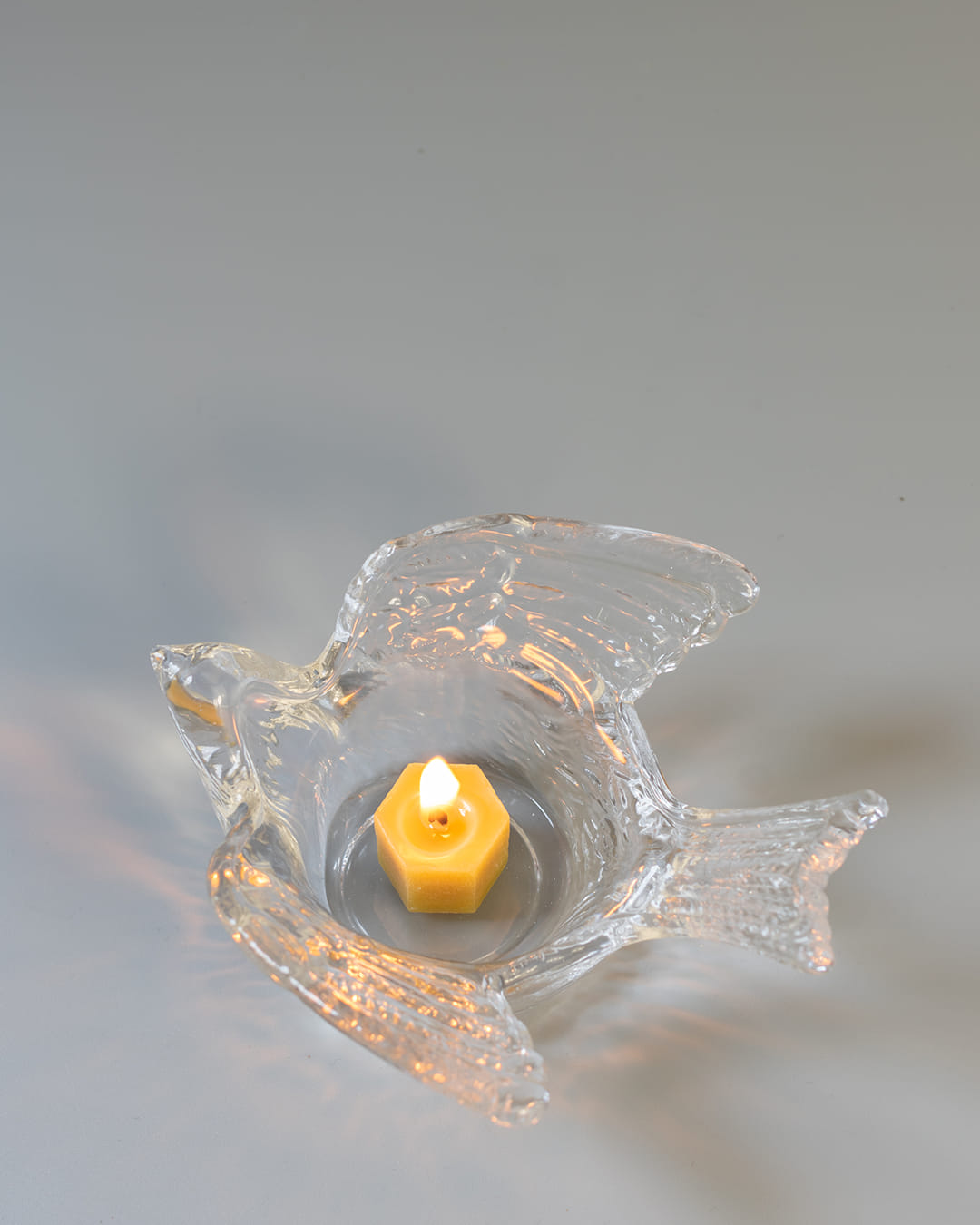 A clear, textured glass tealight holder shaped like a swallow, with a lit yellow tealight candle casting soft light patterns around it.