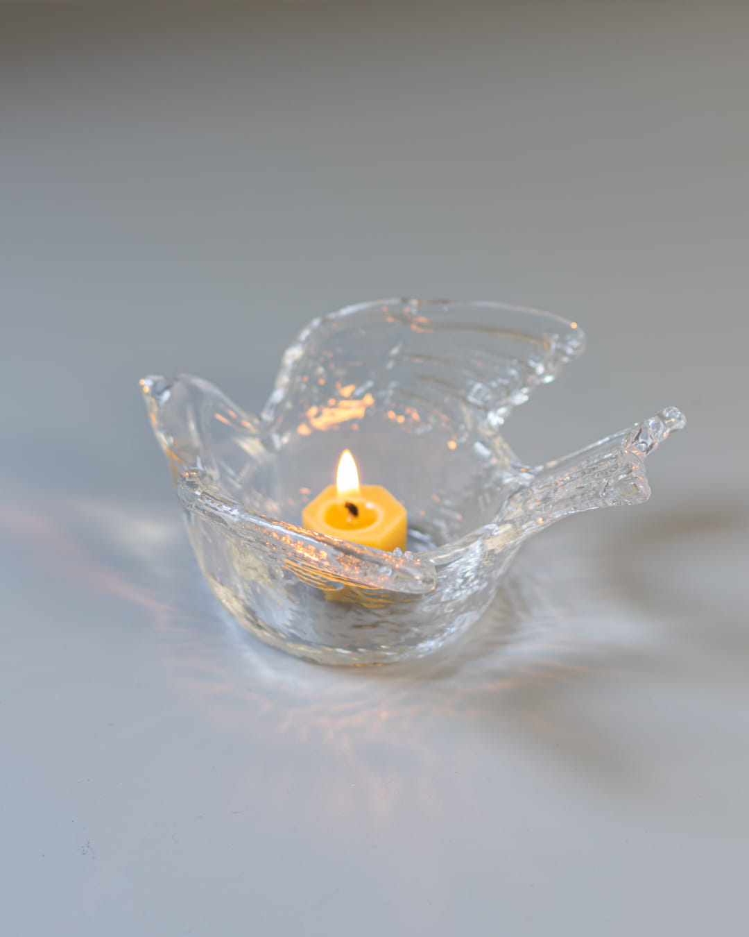 A clear, textured glass tealight holder shaped like a swallow, with a lit yellow tealight candle casting soft light patterns around it.