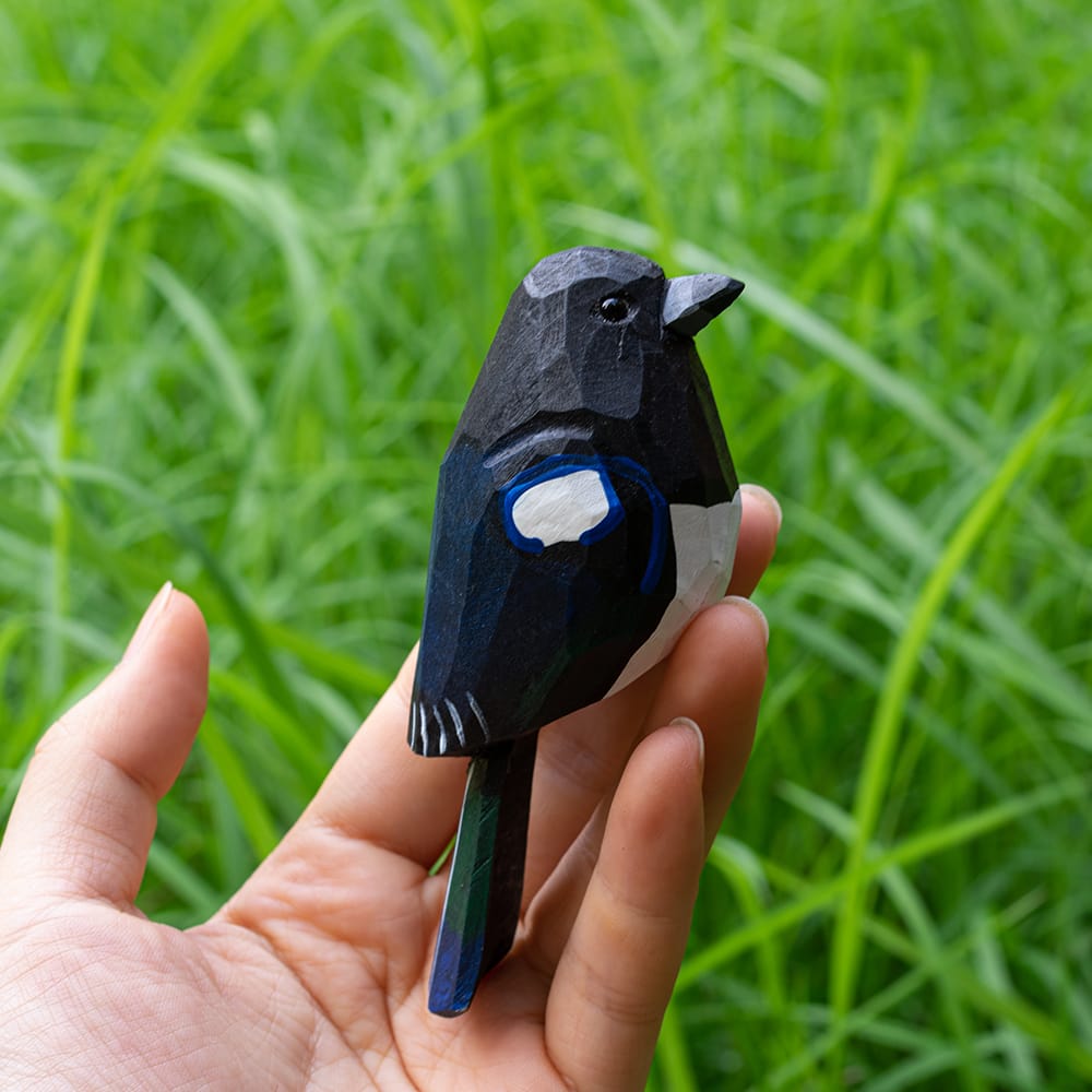 Hand holding a small wooden magpie figurine against a green grass background