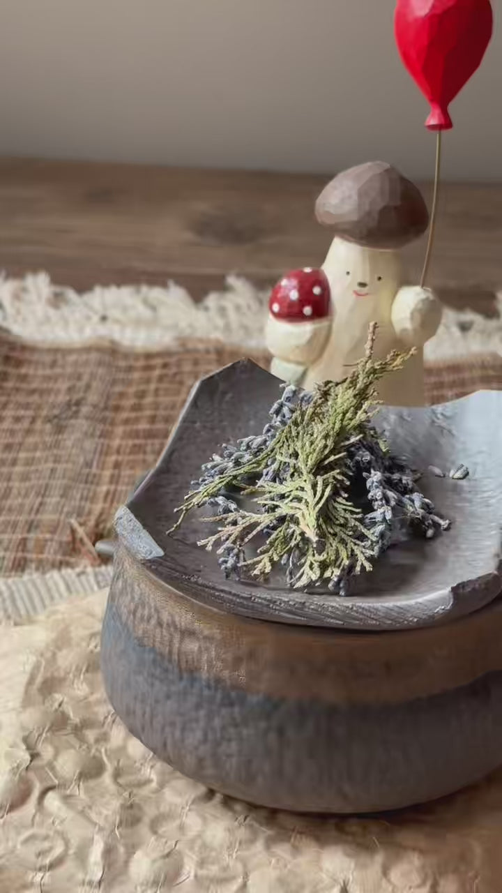 A dark, stone-textured ceramic fragrance warmer (Kawa) with a lit tealight inside, topped with decorative items in the background.